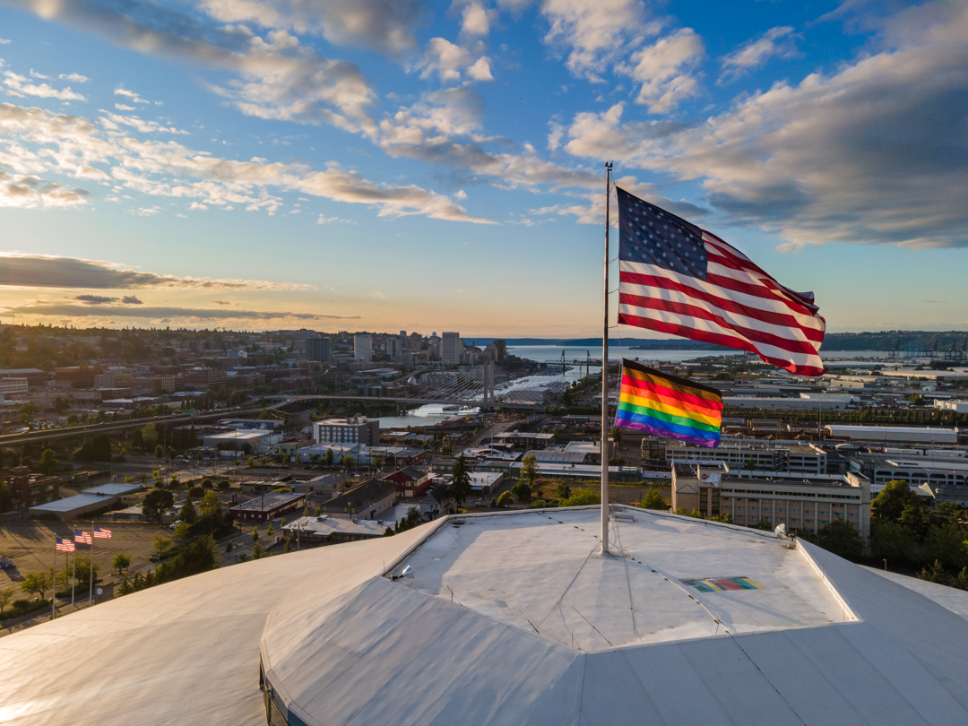 Pride flag and American flag flying above the Tacoma Dome at sunset, with the city and waterfront in the background