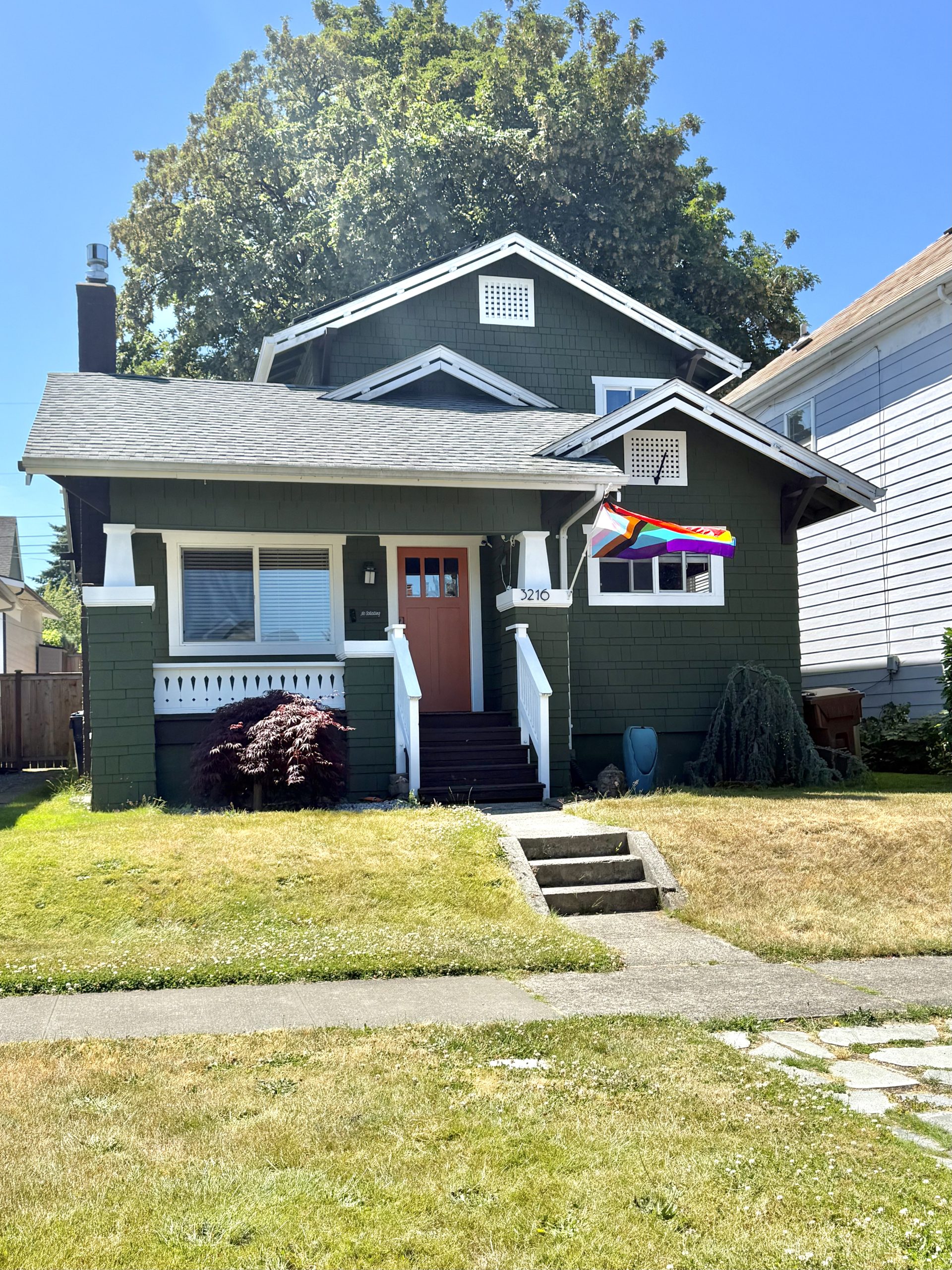 A green Tacoma craftsman bungalow with white trim, a coral-colored front door, and a Progress Pride flag flying from the porch
