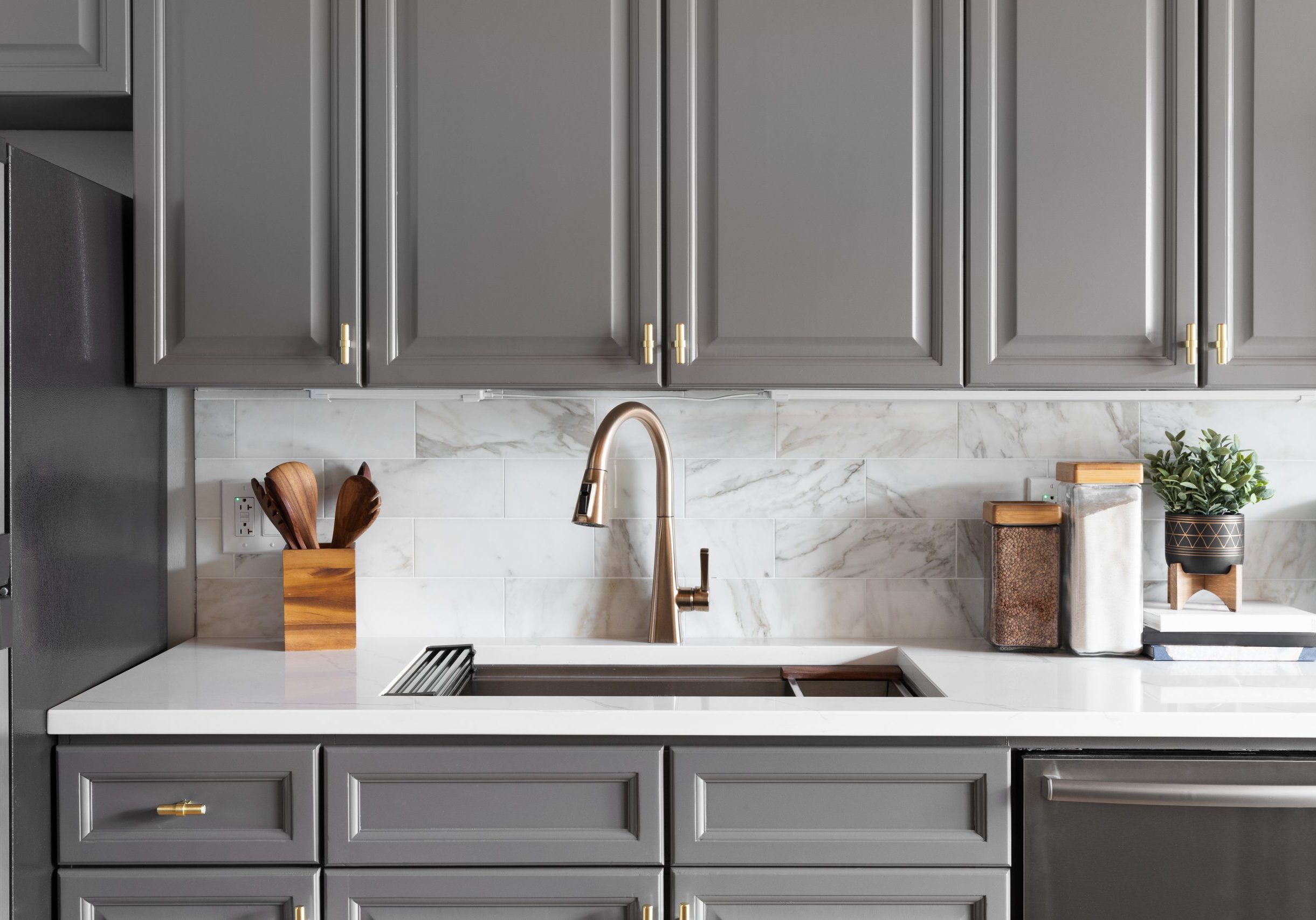 A kitchen sink detail shot with grey cabinets, a white marble co